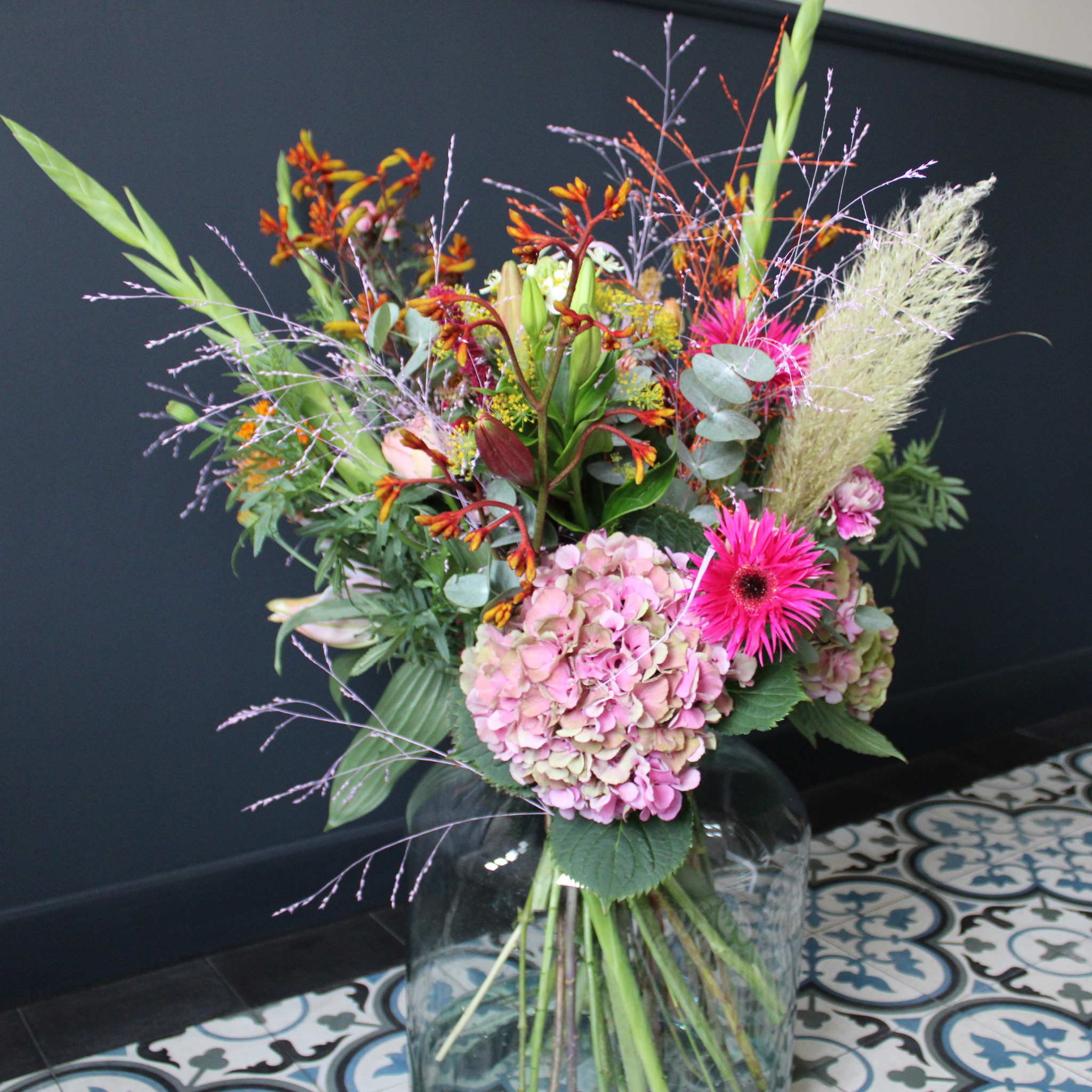 Groot kleurrijk seizoensboeket in glazen vaas voor een donkerblauwe muur – dagverse bloemen van Boeketje van Drosje.